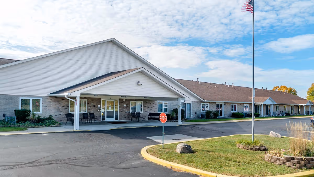 Exterior view of a single-story senior living facility building with a covered entrance, several windows, a stop sign, an American flag on a flagpole, and a landscaped area with grass and rocks under a partly cloudy sky.