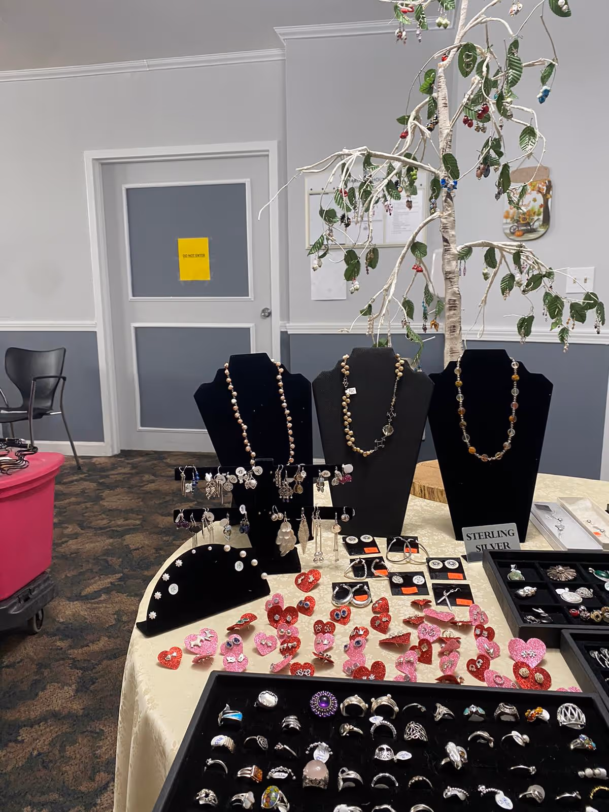 A display table inside a room featuring various jewelry items including rings, necklaces, earrings, and decorative heart-shaped pieces. The jewelry is arranged on black stands and trays on a cream-colored tablecloth. In the background, there is a door with a yellow sign and a wall with some papers pinned on it. The room has gray and white walls and a patterned carpeted floor.