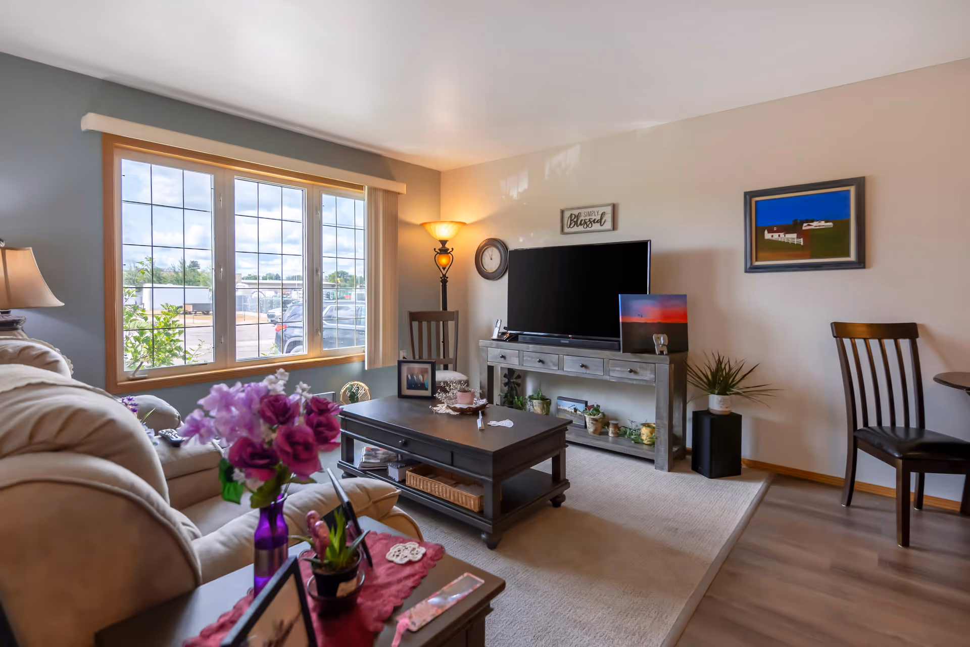 Cozy living room with a sofa, coffee table, TV on a console, and a large window letting in daylight.