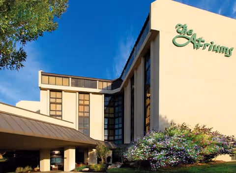 Front exterior of The Atriums senior living building showing its sign, entrance canopy, and landscaped grounds under a blue sky.