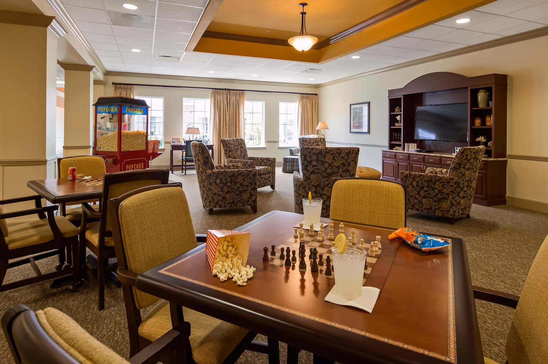 A cozy common area in a senior living facility featuring a table with a chessboard, two glasses of lemonade with lemon slices, a spilled popcorn container, and a bag of chips. In the background, there are several upholstered armchairs arranged around a large wooden entertainment center with a flat-screen TV. A popcorn machine is visible near the windows, which let in natural light through beige curtains.