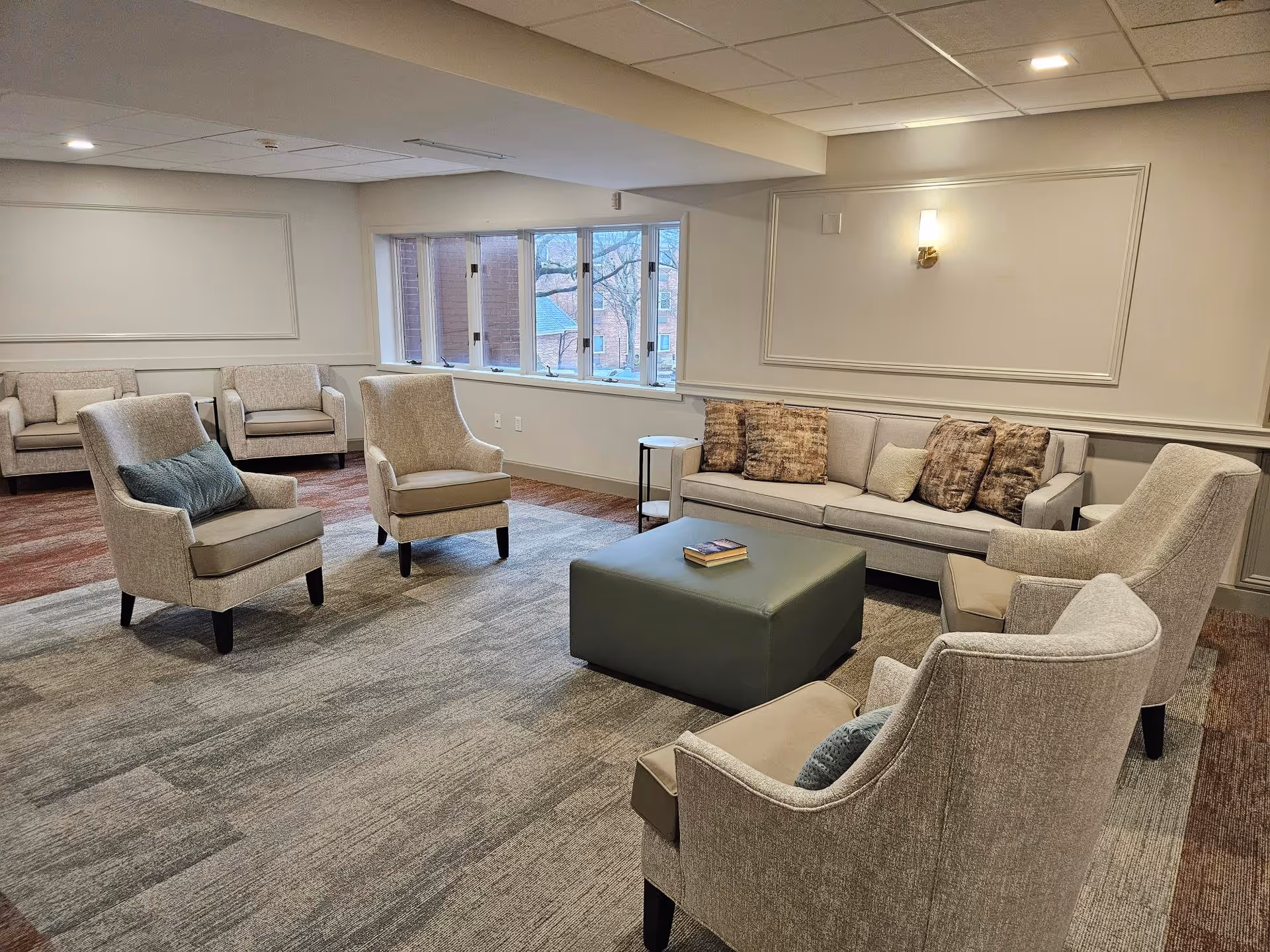 A spacious senior living common area with beige upholstered armchairs and a matching sofa adorned with patterned cushions. A large green ottoman with a book on top is centered on a gray carpet. The room has a window showing an outside view of trees and buildings, neutral-colored walls with decorative molding, and a wall-mounted light fixture.