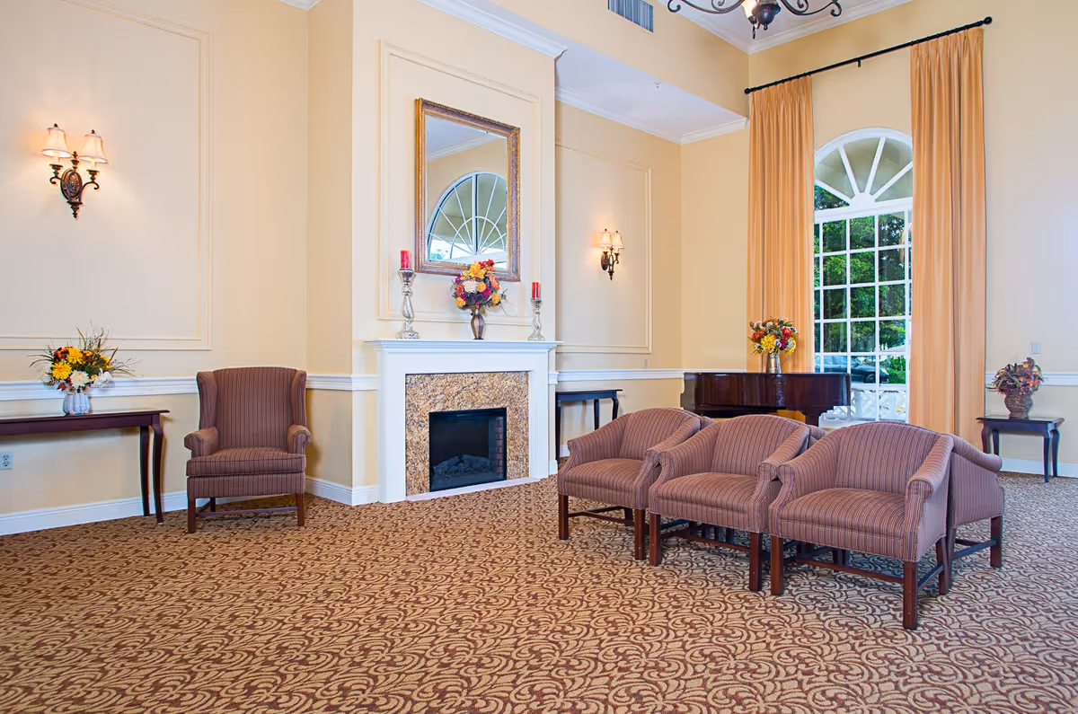 A spacious and elegant living room area with patterned carpet, three striped armchairs, a single armchair, a white fireplace with a mirror above it, wall sconces with lamps, and large windows with long beige curtains. There are flower arrangements on tables and a grand piano near the window.