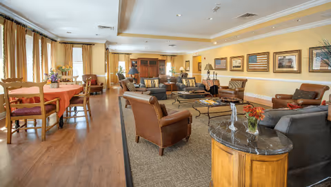 Spacious senior living common room with sofas, leather armchairs, coffee tables and a dining table by windows.
