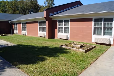 Single-story red-brick facility exterior with windows, a green lawn, and a small raised garden bed.
