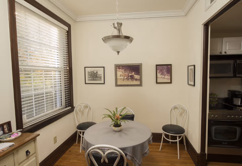 A small dining area with a round table covered with a gray tablecloth and a potted plant centerpiece. Four white chairs with black cushions surround the table. The room has light-colored walls with three framed pictures of horse racing on one wall. A window with white blinds is on the left side, and a hanging light fixture is above the table. Part of a kitchen with a stove and microwave is visible through an open doorway on the right.