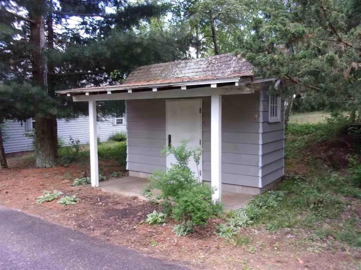 A small gray shed with a white door and a shingled roof, surrounded by trees and greenery. There is a small concrete porch with two white support posts and some plants growing in front of the shed. The area around the shed is covered with mulch and bordered by a paved path.