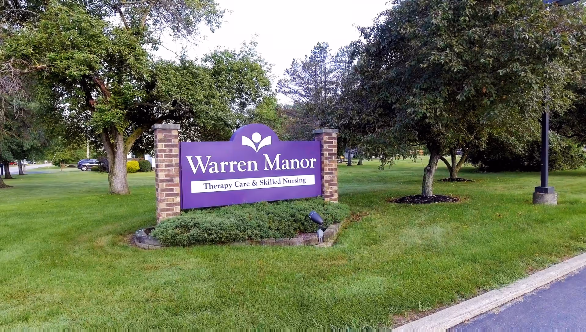 Outdoor view of a green lawn with trees and a purple sign reading 'Warren Manor Therapy Care & Skilled Nursing' supported by two brick pillars.