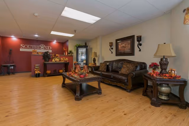 A cozy living room area in Southland Rehabilitation And Healthcare Center featuring a dark brown leather sofa with decorative pillows, a wooden coffee table with autumn-themed decorations, and matching side tables with lamps and seasonal decor. The background shows a red accent wall with the Southland sign and additional decorative items.