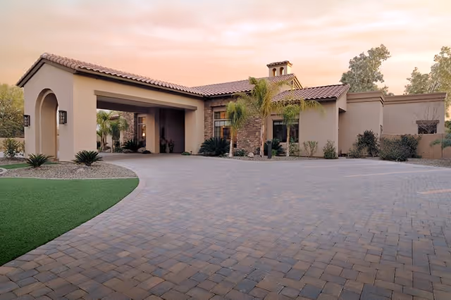 Exterior view of a single-story building with a tiled roof and stone accents, featuring a covered entrance and surrounded by landscaped plants and palm trees under a soft evening sky.