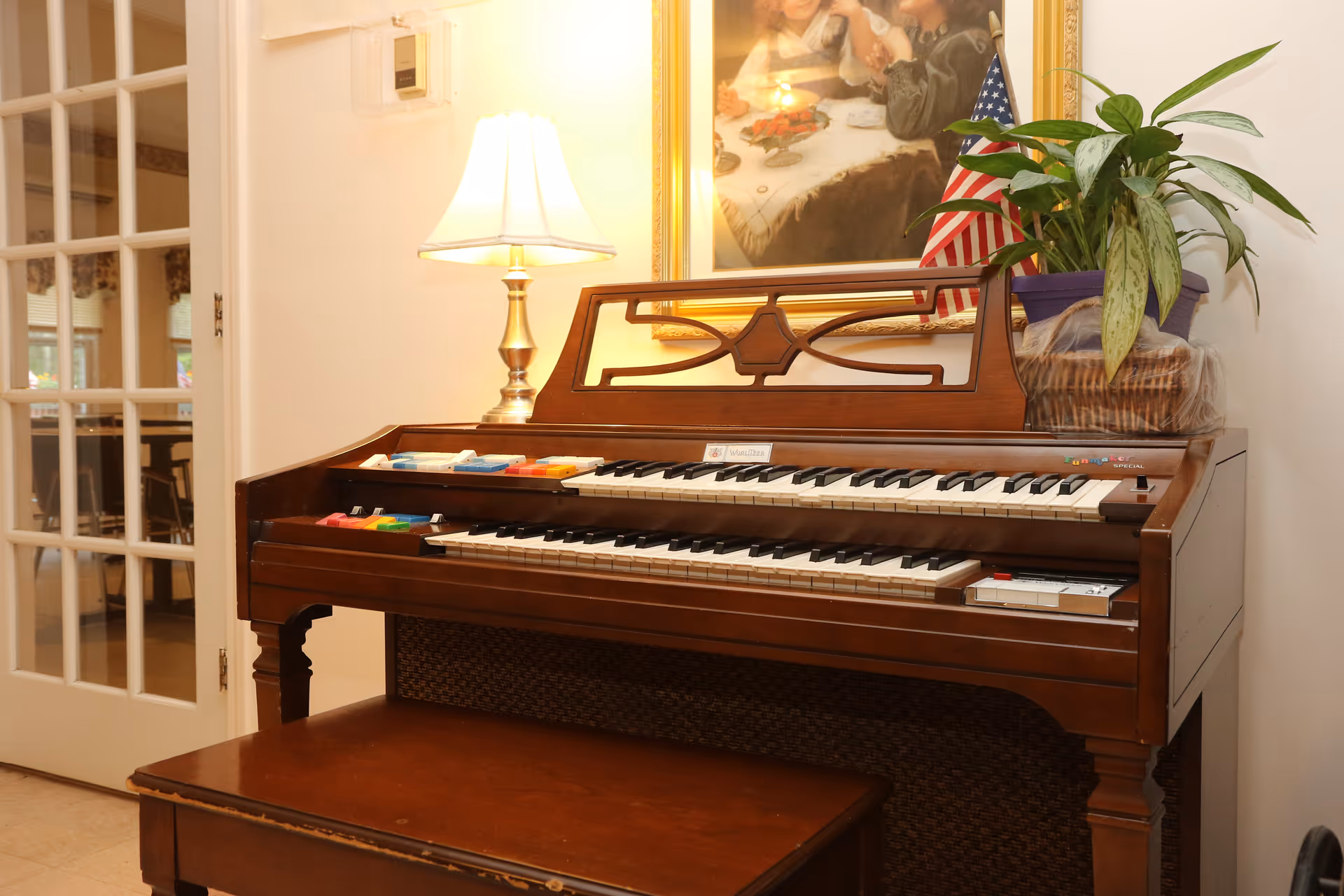 A wooden organ with two keyboards and colorful switches on top, a matching wooden bench in front, a table lamp with a white shade on the left side, a potted plant and a small American flag on top of the organ, and a framed painting of children in the background. A glass-paneled door is visible to the left.