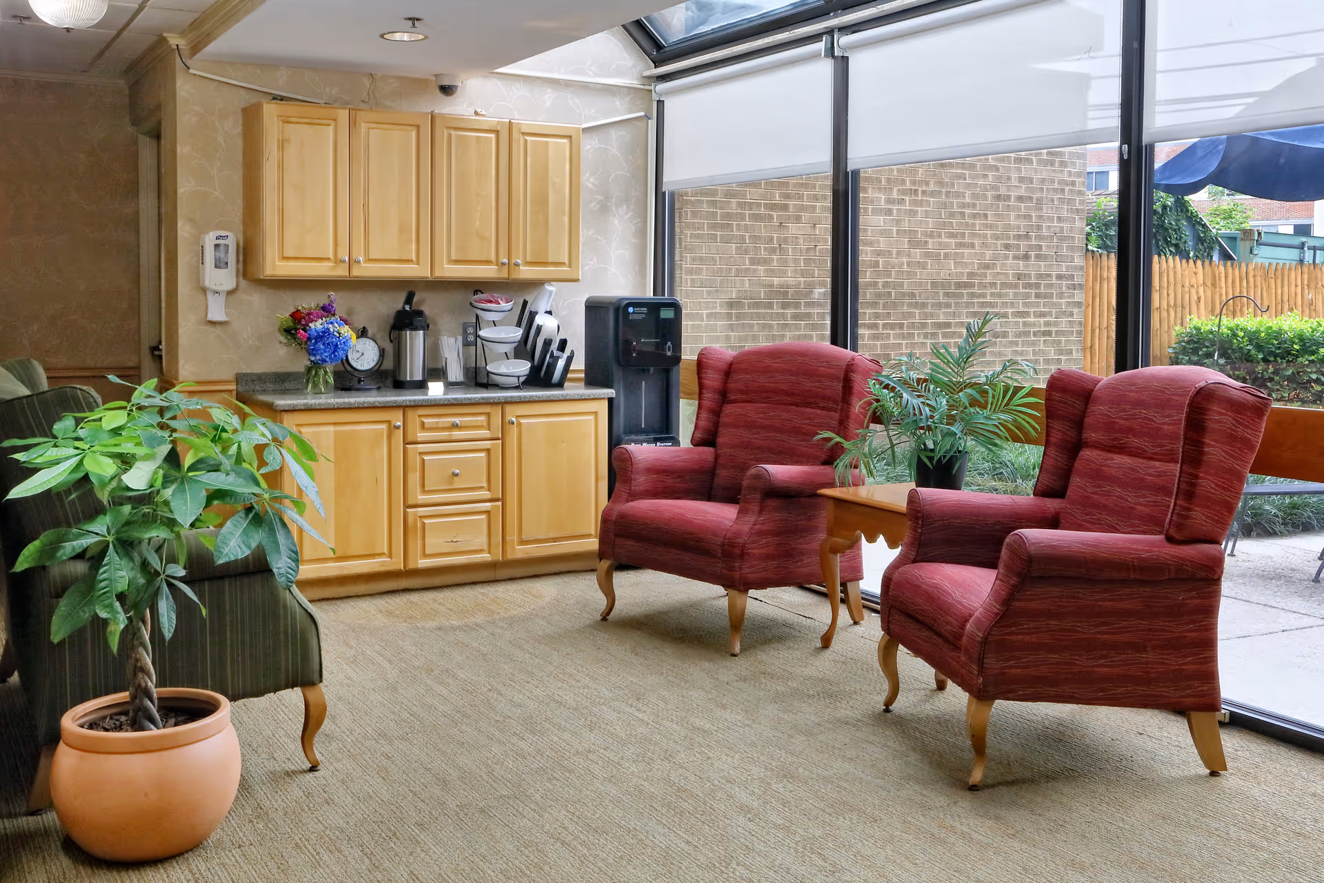 Sunlit common room with two red armchairs, a small wooden table, potted plants, and a kitchenette/coffee station along a wall of windows.