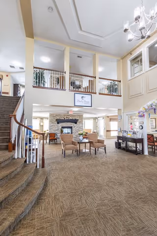 Spacious and well-lit senior living facility common area with a carpeted floor, a curved staircase with wooden handrails on the left, and a seating area with four cushioned chairs and a small table in front of a stone fireplace. The room has high ceilings with a chandelier, large windows, and a balcony railing overlooking the area. A sign reading 'Bailey Pointe' is mounted above the fireplace.