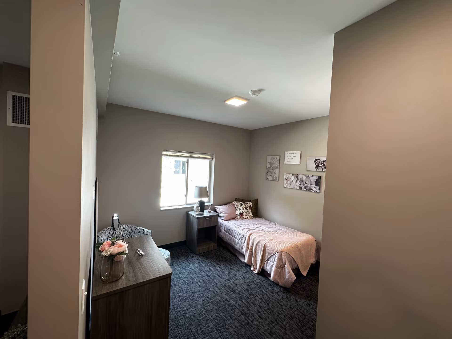 A small, cozy bedroom with a single bed covered in a pink blanket and decorative pillows. There is a nightstand with a lamp and a clock next to the bed. A window lets in natural light. On the opposite side, there is a wooden dresser with a vase of flowers and a small mirror on top. The walls are decorated with framed artwork and quotes. The room has carpeted flooring and neutral-colored walls.