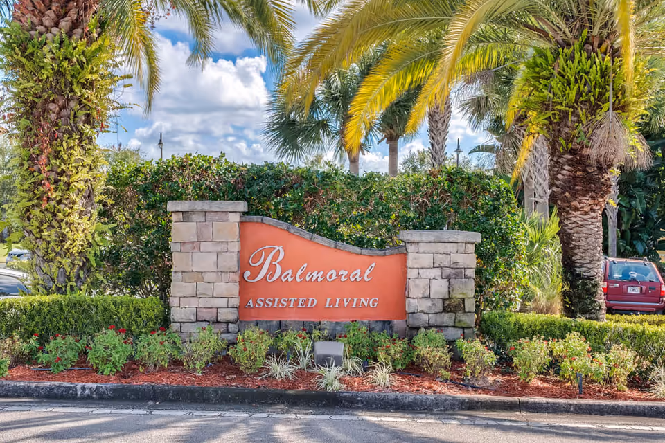 Stone and brick sign for Balmoral Assisted Living surrounded by landscaped bushes and palm trees under a partly cloudy sky.