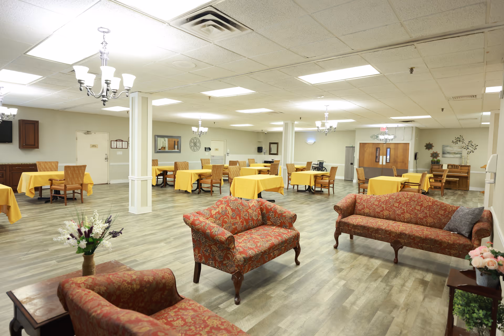 A spacious room with several tables covered in yellow tablecloths and wooden chairs arranged around them. In the foreground, there are three upholstered sofas with a red and gold floral pattern, a small wooden table with a vase of flowers, and a wooden side table with a plant and flowers. The room has a light-colored wood floor, white walls, and multiple chandeliers hanging from the ceiling. There are doors and wall decorations visible in the background.