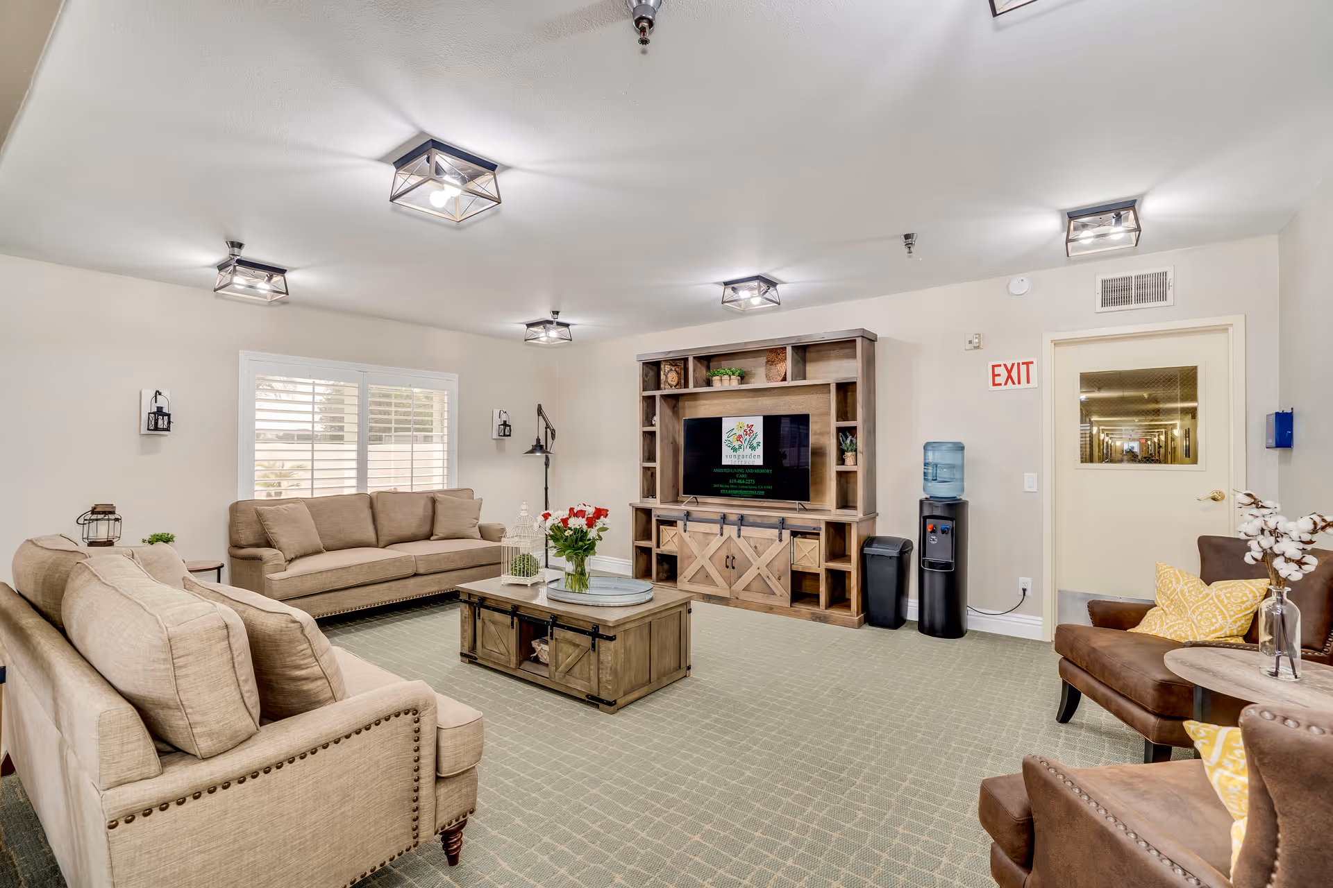 Well-lit assisted living common room with beige sofas and armchairs arranged around a coffee table facing a wooden entertainment center and TV.