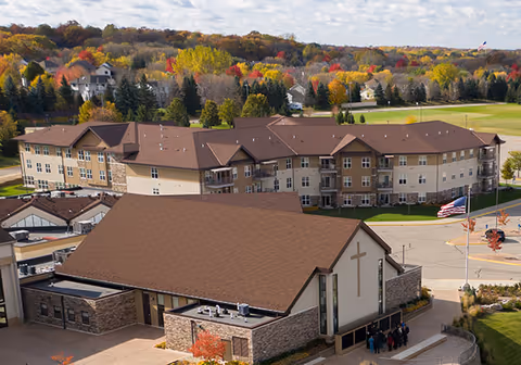 Aerial view of The Fountains at Hosanna senior living facility showing a large multi-story building with beige and brown exterior surrounded by trees with autumn foliage. In the foreground, there is a smaller building with a cross on the front, likely a chapel, and an American flag flying nearby. A group of people is gathered near the entrance of the smaller building.