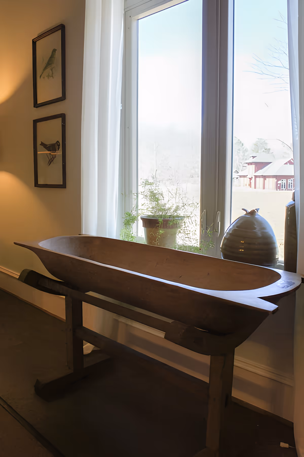 A wooden decorative trough or bowl on a stand placed near a window with white curtains. Outside the window, there is a view of a field and a red barn. Two framed bird illustrations hang on the wall to the left.