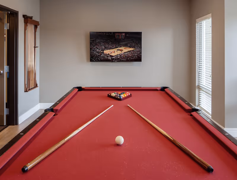 A red pool table with two pool cues and a rack of billiard balls set up for a game in a room with beige walls. A flat-screen TV mounted on the wall displays a basketball game. There is a window with blinds on the right side and a wooden cue rack on the left wall.