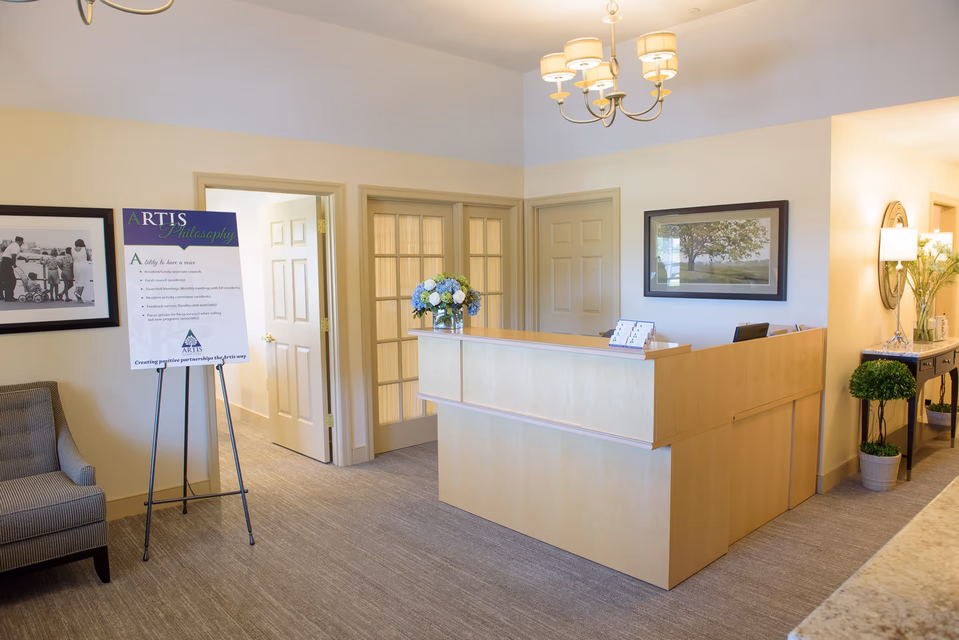 Reception area with a light wood front desk adorned with a vase of blue and white flowers. To the left, there is a gray upholstered chair and a sign on an easel titled 'ARTIS Philosophy'. The walls are light-colored with framed pictures, and there is a chandelier hanging from the ceiling. A hallway and several doors are visible in the background.