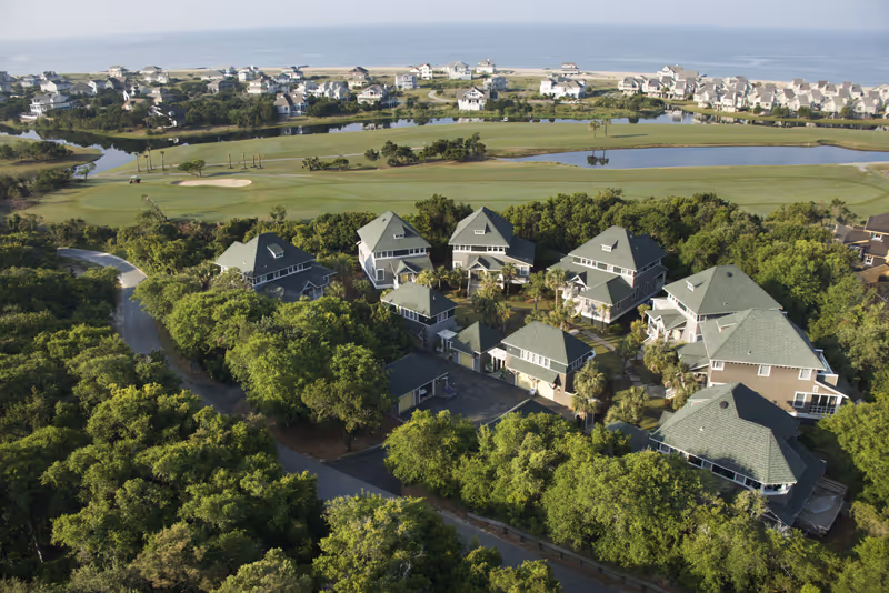 Aerial view of a residential community with multiple houses surrounded by trees, a golf course with sand traps and water hazards, and the ocean in the background.