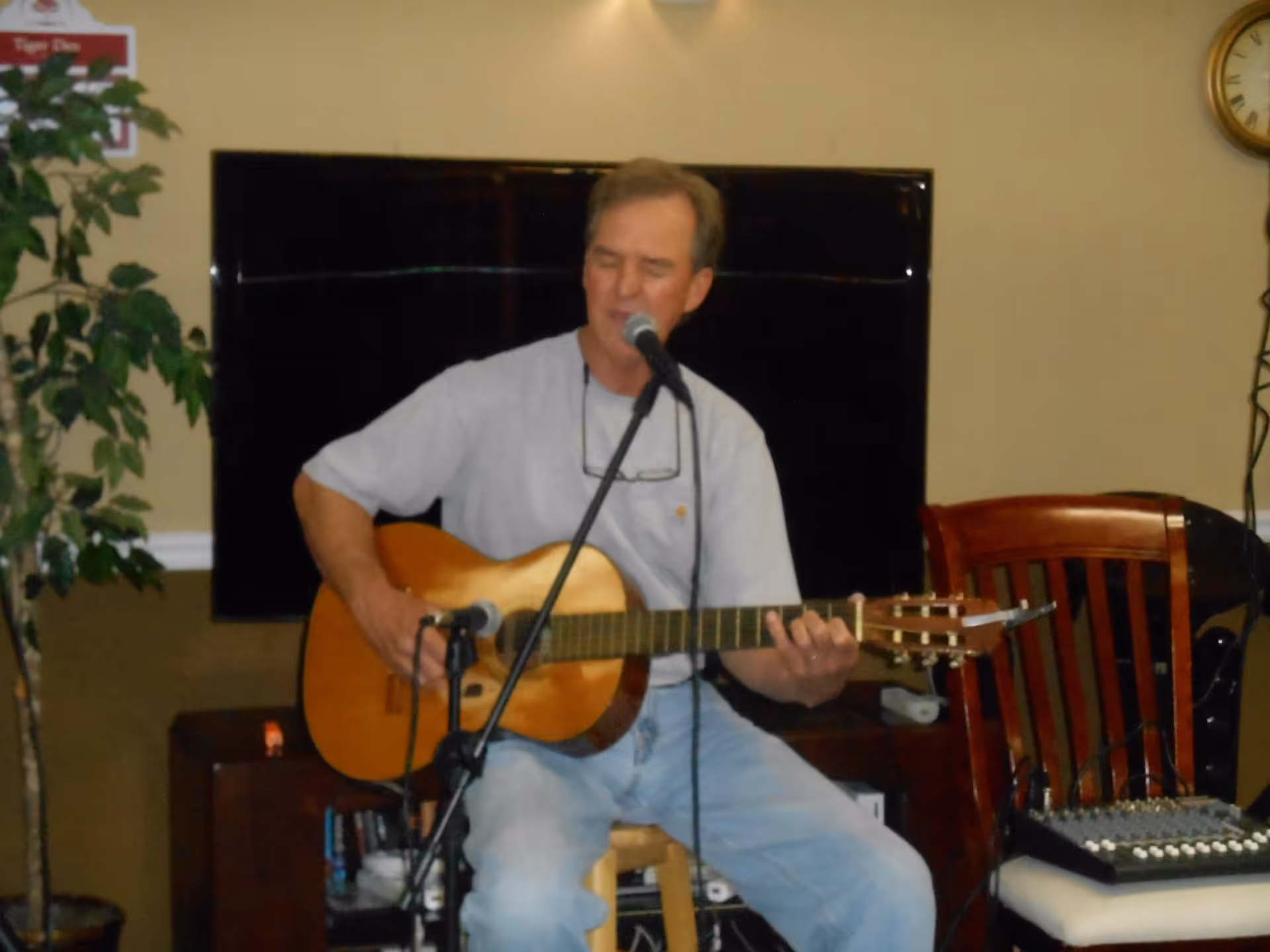 A man sitting on a stool playing an acoustic guitar and singing into a microphone in a room with a large flat-screen TV mounted on the wall behind him. There is a wooden chair with audio equipment on it to his right and a potted plant to his left.