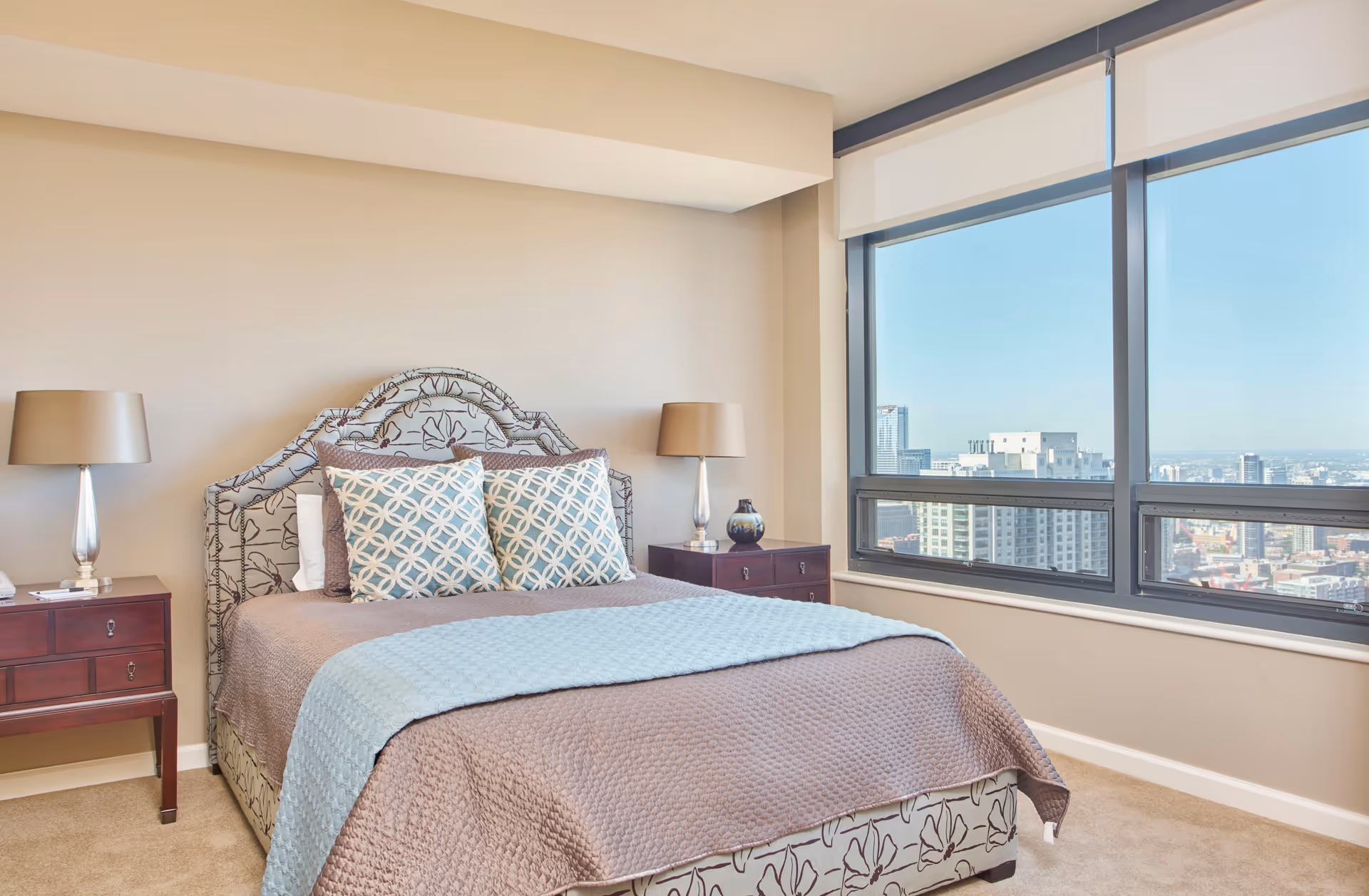 A neatly made bed with a patterned headboard and two decorative pillows in a bedroom. There are two bedside tables with matching lamps on either side of the bed. A large window offers a view of a cityscape with tall buildings under a clear blue sky.
