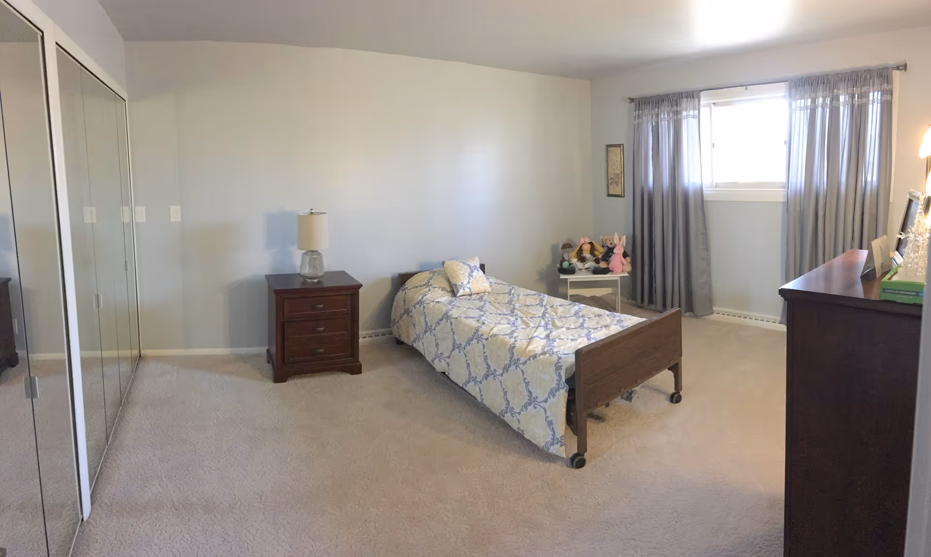 A simple bedroom with a single bed covered in a patterned bedspread, a wooden nightstand with a lamp, a small table with stuffed animals, a window with gray curtains, a wooden dresser, and mirrored closet doors.