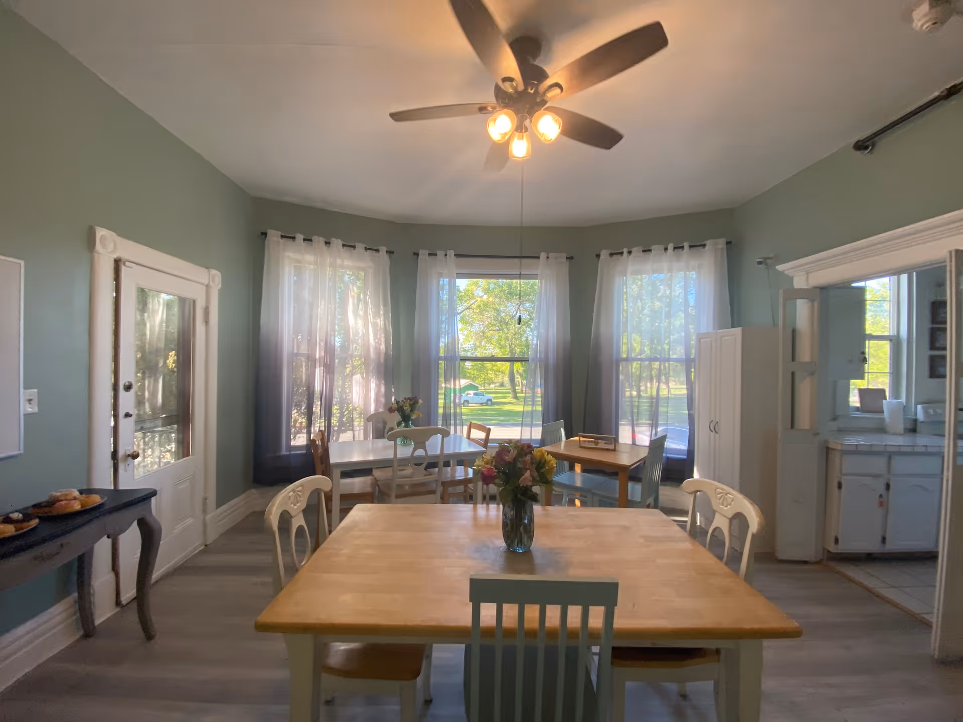 Bright dining area with wooden tables and chairs, a ceiling fan, large bay windows with sheer curtains, and a vase of flowers on the nearest table.