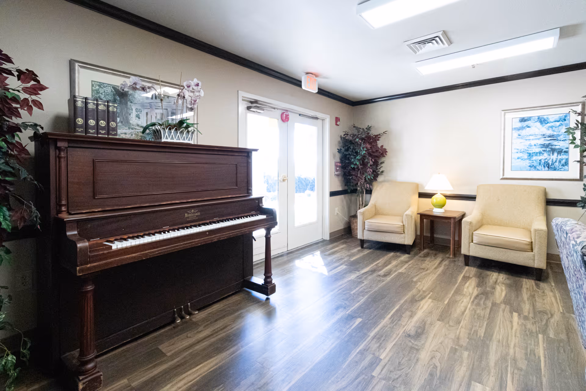 A bright common room with an upright piano on the left, double doors in the center, and two beige armchairs with a side table and lamp on the right.