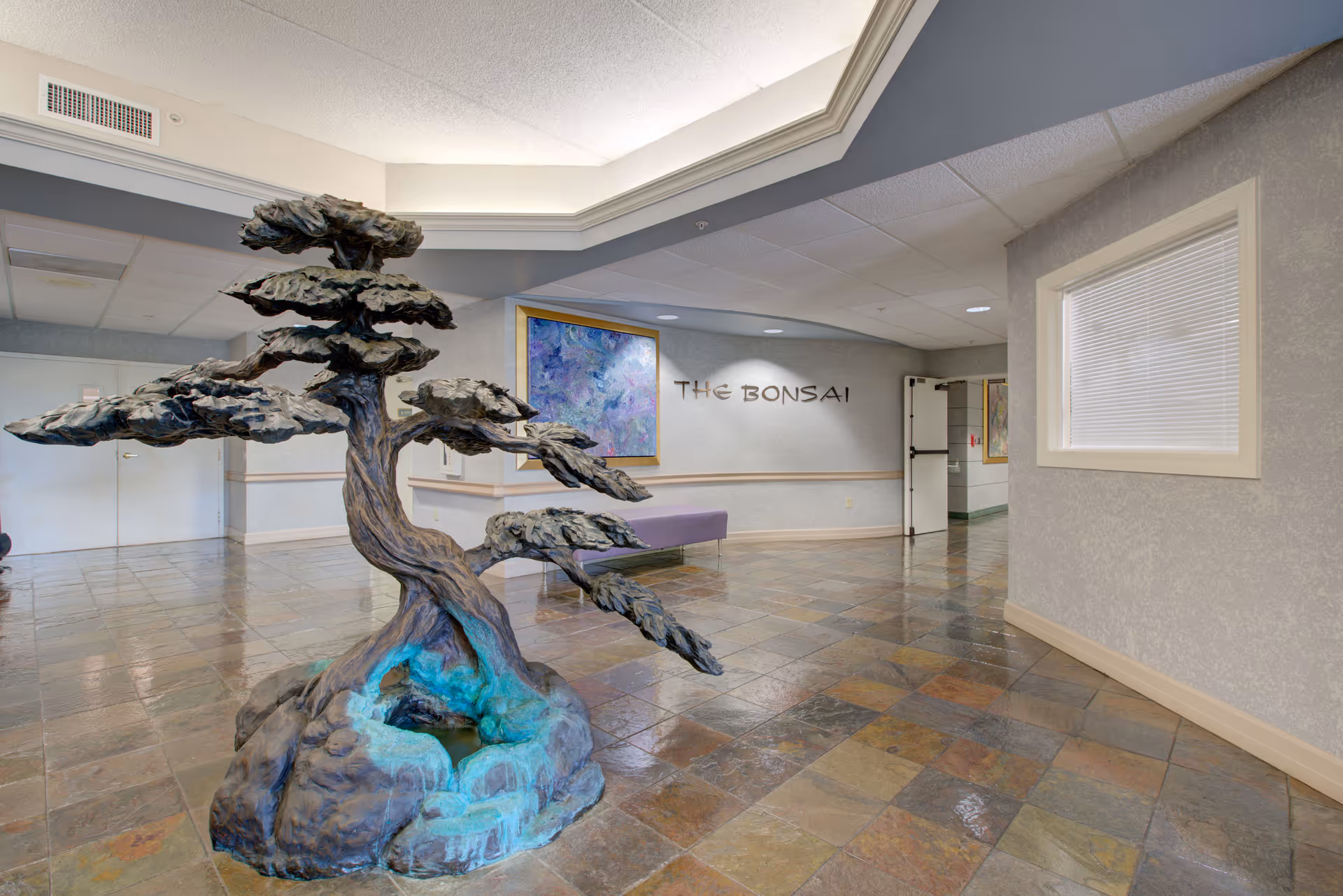 Interior view of a senior living facility hallway featuring a large bonsai tree sculpture on a stone base in the center. The floor is tiled with multicolored stone tiles, and there is a purple bench against the wall. A framed abstract painting hangs on the wall above the bench, and the words 'THE BONSAI' are displayed on the wall. The ceiling has recessed lighting and a decorative tray ceiling.