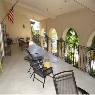 Covered outdoor balcony area with metal chairs and tables, decorative iron railing, arched openings overlooking greenery, and an American flag mounted on the wall.