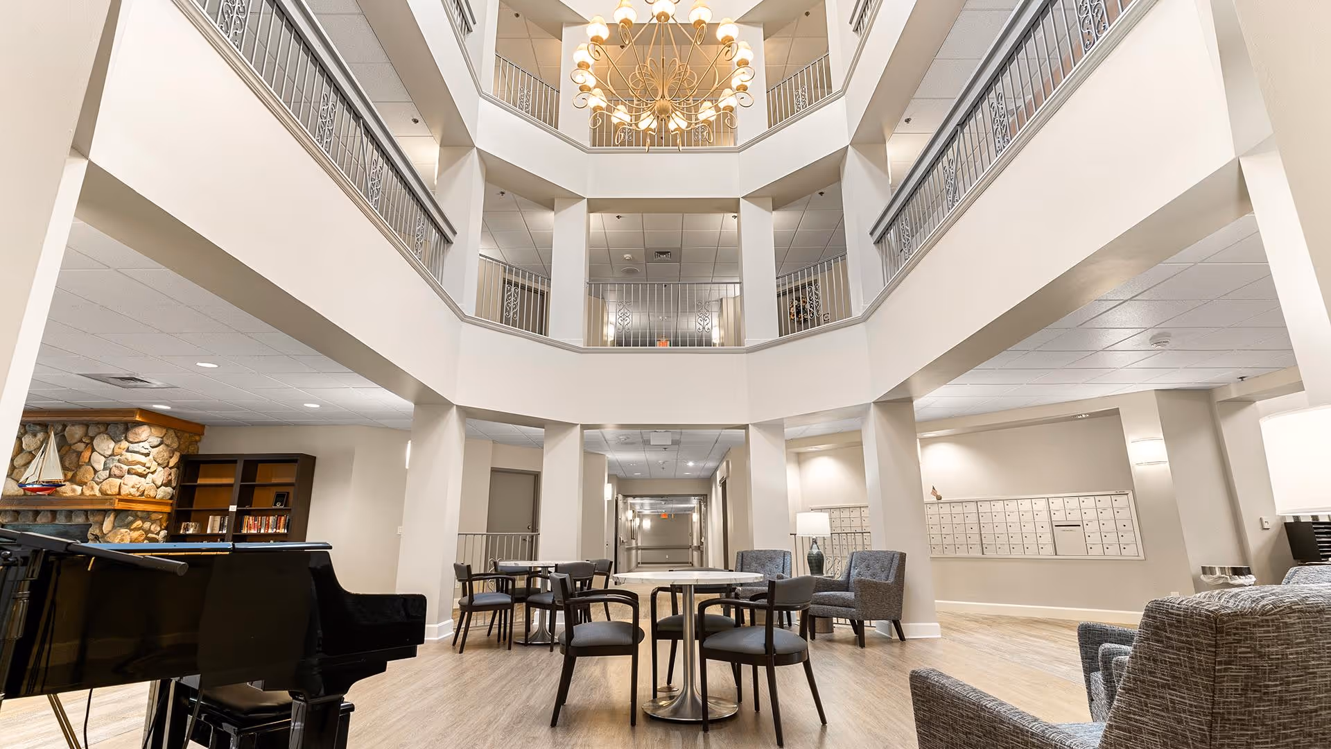 A spacious, multi-level common area inside a senior living facility with a grand chandelier hanging from the ceiling. The area features a black grand piano, several round tables with chairs, comfortable armchairs, a stone fireplace, bookshelves, and a wall of mailboxes. The space is well-lit with neutral-colored walls and wood flooring.