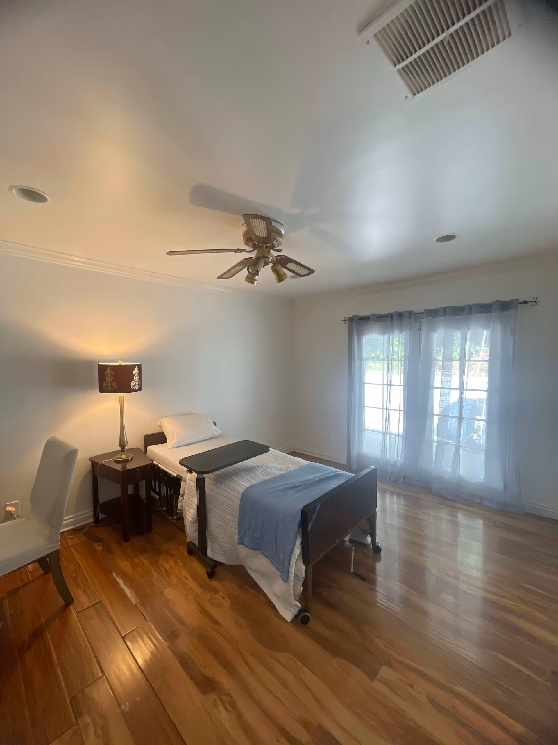 A simple bedroom with a single hospital-style bed covered with white and blue bedding. Next to the bed is a small wooden nightstand with a decorative lamp. A white chair is positioned near the nightstand. The room has wooden flooring, white walls, a ceiling fan with lights, and large windows with sheer curtains allowing natural light to enter.
