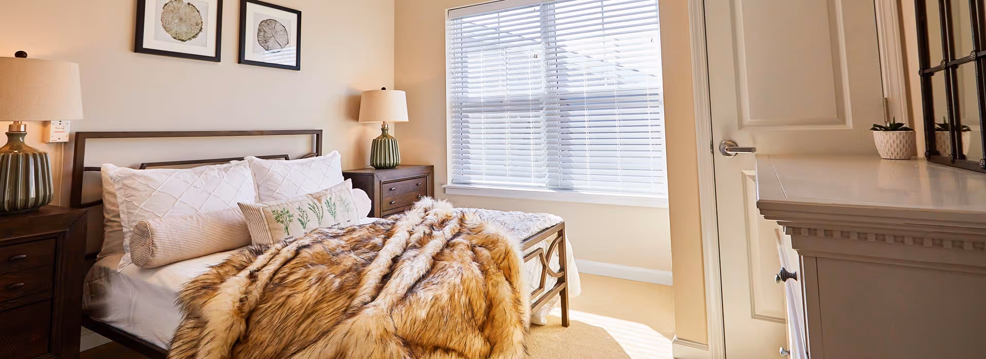 Bright furnished bedroom with a bed draped in a fur throw, nightstands with lamps, a dresser, and a window with blinds.