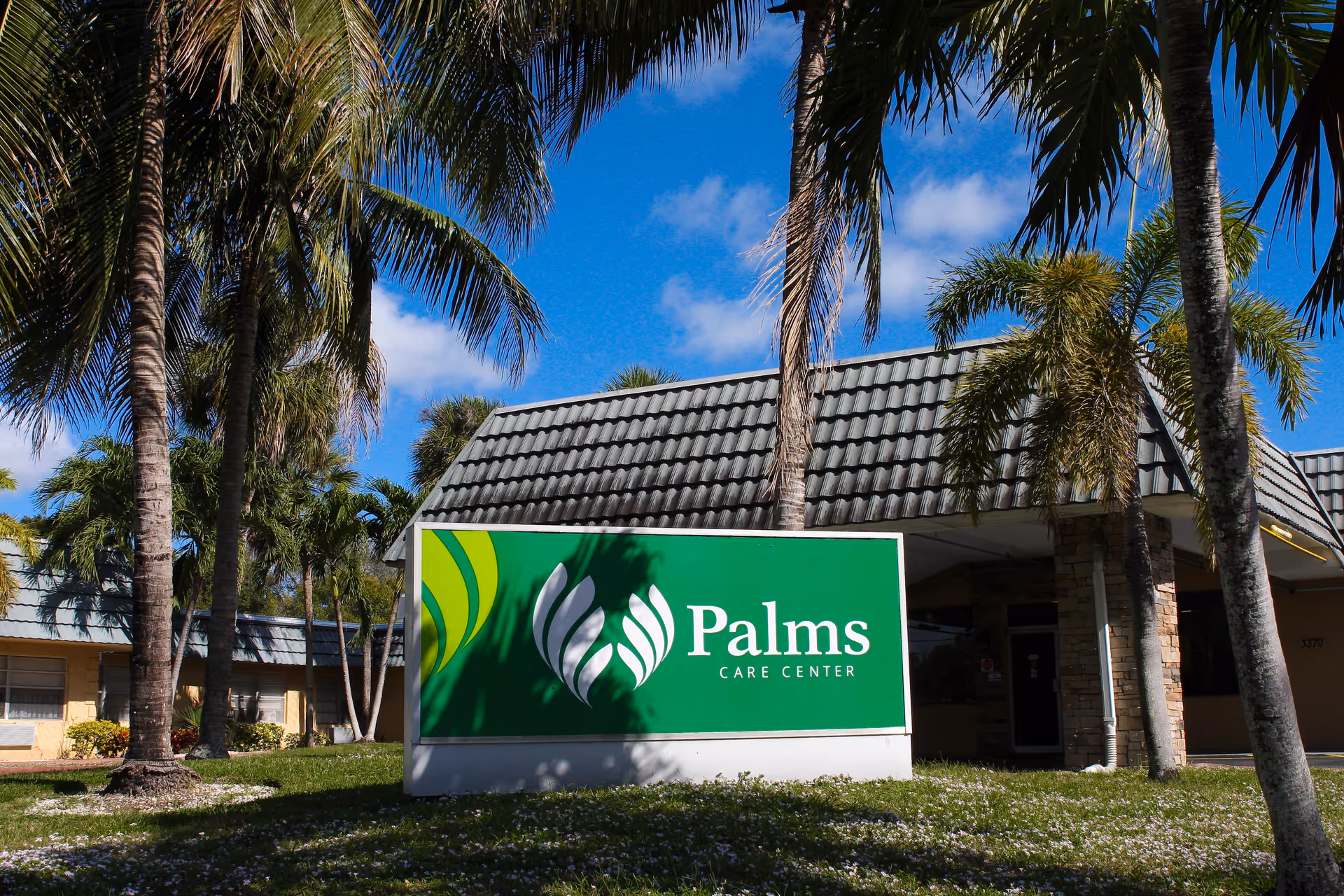 A green 'Palms Care Center' sign sits on a lawn in front of a tiled-roof building surrounded by palm trees under a blue sky.