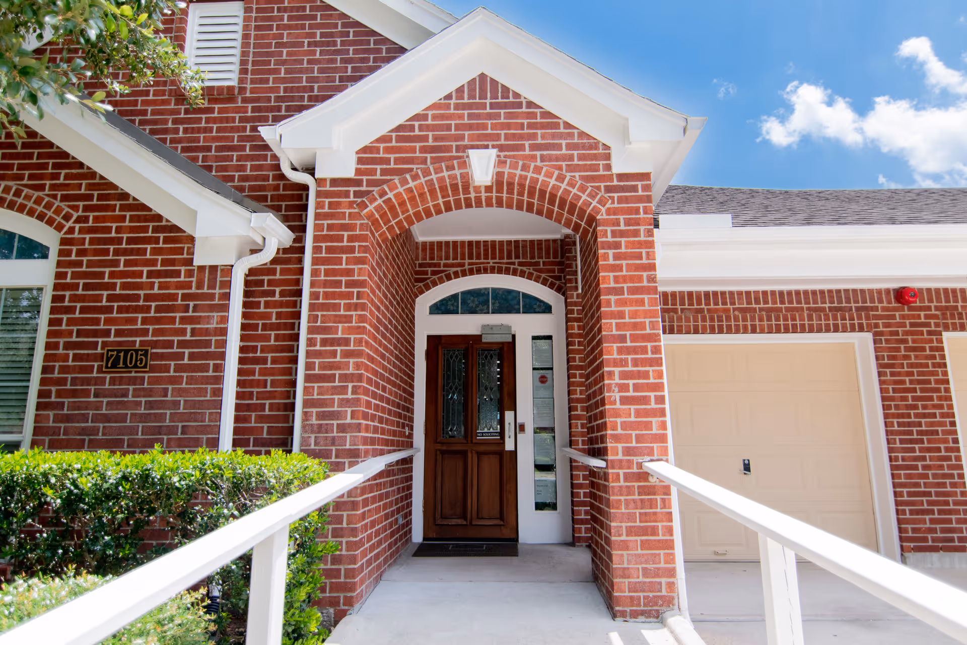 Entrance of a brick building with a wooden door under an arched brick entryway, white railings on both sides of the ramp leading to the door, a garage door to the right, and green bushes on the left under a blue sky with some clouds.
