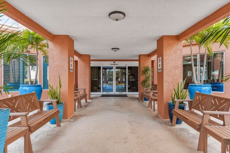 Covered entrance area of a building with brown benches and blue plant pots containing green plants on either side. The entrance has automatic sliding glass doors and orange textured pillars supporting the ceiling.