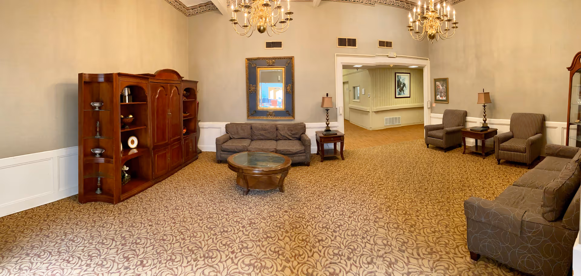 Spacious lounge with sofas and armchairs around a central coffee table, wooden shelving, chandeliers, and patterned carpet.