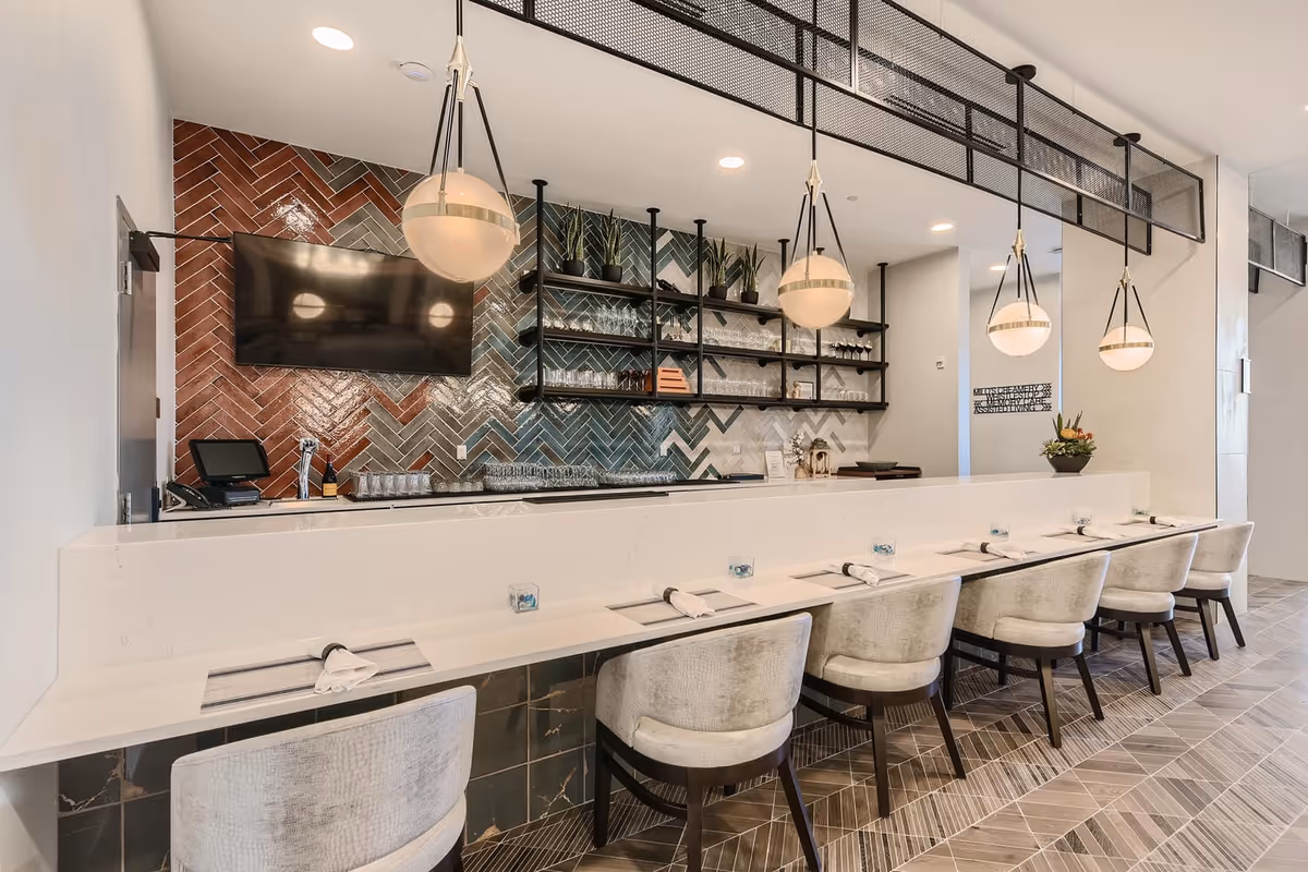 Modern dining area with a long white counter and eight cushioned chairs. Above the counter hang four spherical pendant lights. Behind the counter is a wall with a colorful herringbone tile pattern, open black shelving with glassware and plants, and a mounted flat-screen TV.