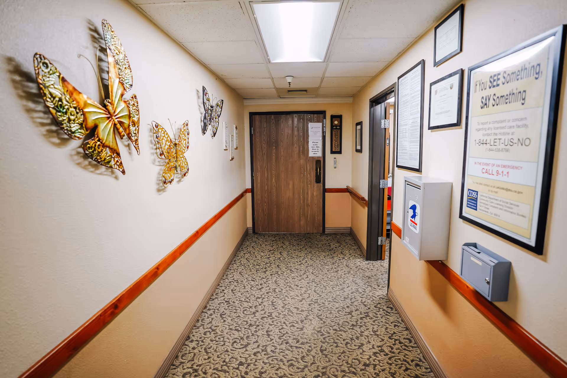 A carpeted hallway in an assisted living facility with decorative metal butterfly wall art on the left wall and framed notices on the right wall. At the end of the hallway is a wooden door with a sign, and there is a handrail running along both walls.