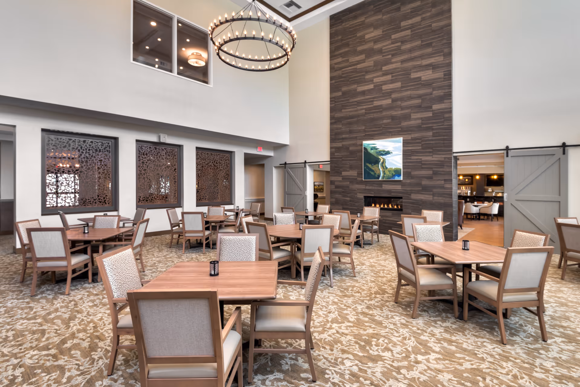 A spacious dining room with multiple wooden tables and cushioned chairs arranged neatly on a patterned carpet. The room features a tall ceiling with a large circular chandelier, a modern fireplace set in a dark textured wall, and a landscape painting above the fireplace. There are decorative window panels on one wall and sliding barn doors leading to another room.
