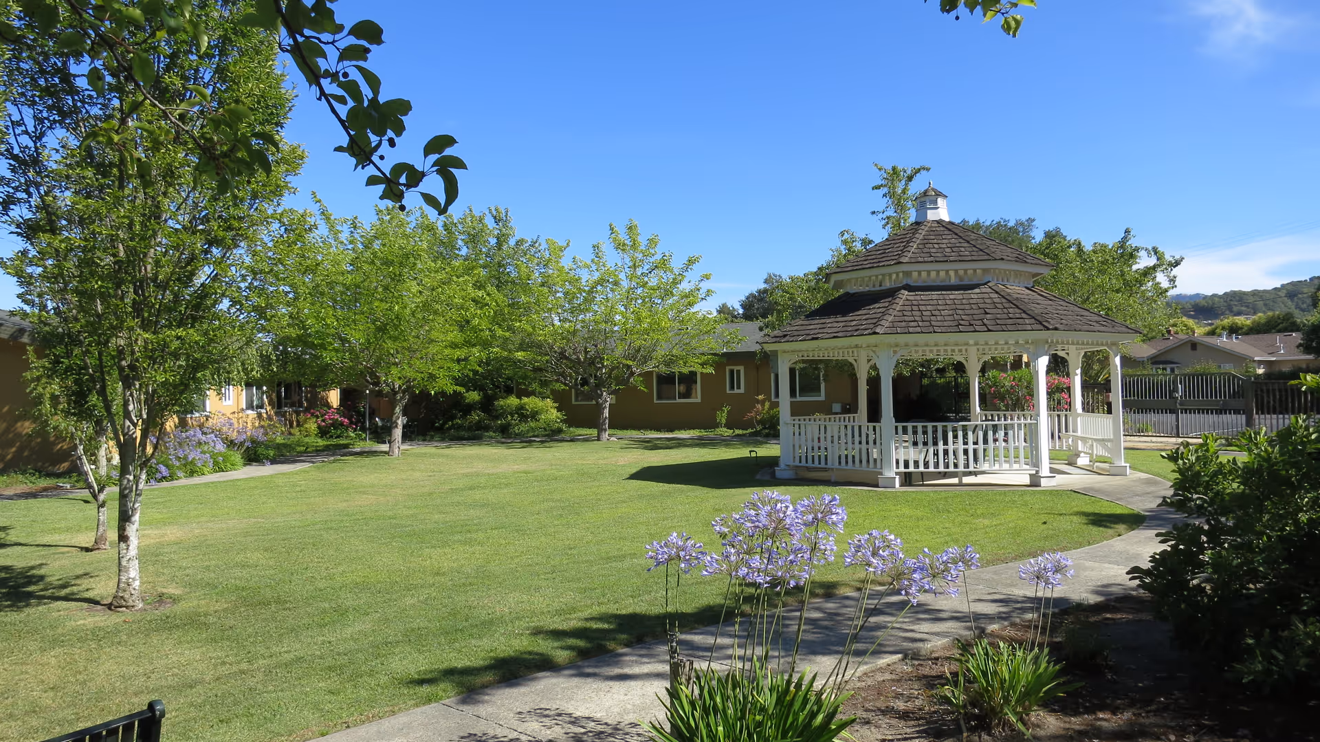 A well-maintained outdoor garden area at Creekwood Care Facility featuring a white wooden gazebo with a shingled roof, surrounded by green grass, trees, and flowering plants under a clear blue sky.