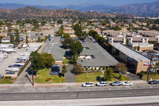 Aerial view of a single-story care center building with a gray roof, surrounded by parking lots and green lawns. Several cars are parked in front and around the building. The facility is located in a suburban area with many houses and hills in the background under a clear sky.