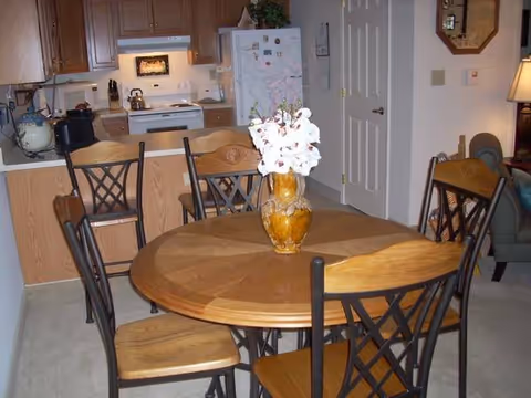 Round wooden dining table with a vase of flowers and chairs in an open-plan room with a kitchen visible in the background.