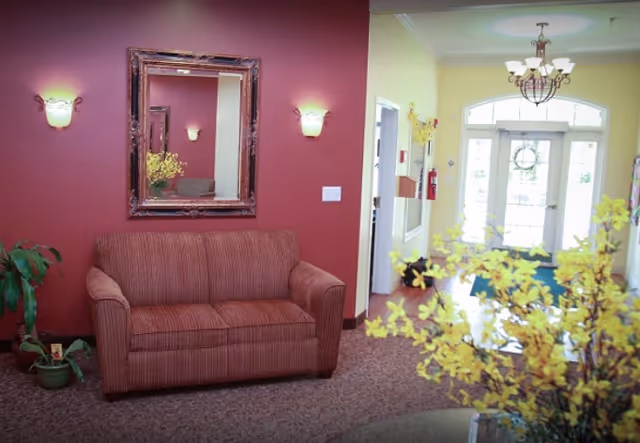 A cozy seating area with a striped red loveseat against a red wall, flanked by two wall sconces and a large framed mirror. To the right, there is a hallway leading to a glass door entrance with a decorative wreath, and a chandelier hanging from the ceiling. Yellow flowers are visible in the foreground.