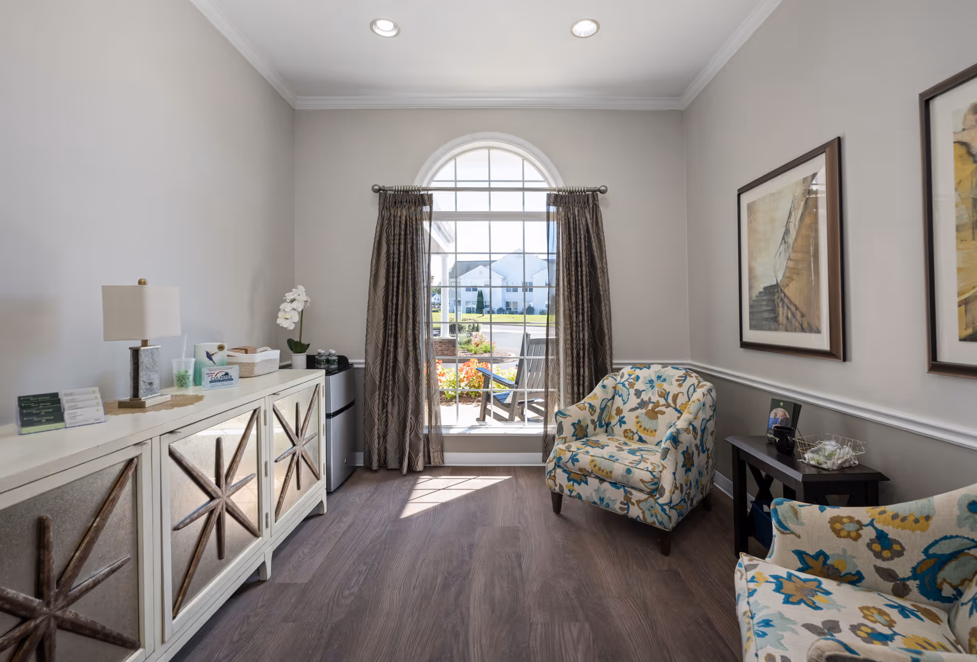 A bright sitting area with two floral patterned armchairs, a small dark wooden side table, and a white cabinet with decorative star designs. A large arched window with brown curtains lets in natural light and offers a view of outdoor chairs and a garden. The walls are painted light gray with white trim, and two framed artworks hang on the wall.