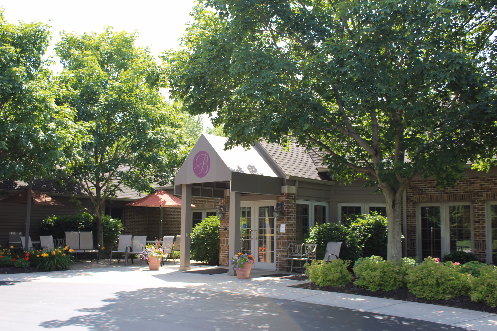 Entrance of Burton's Ridge Assisted Living facility with a covered entryway, surrounded by green trees, shrubs, and outdoor seating areas with chairs and umbrellas.