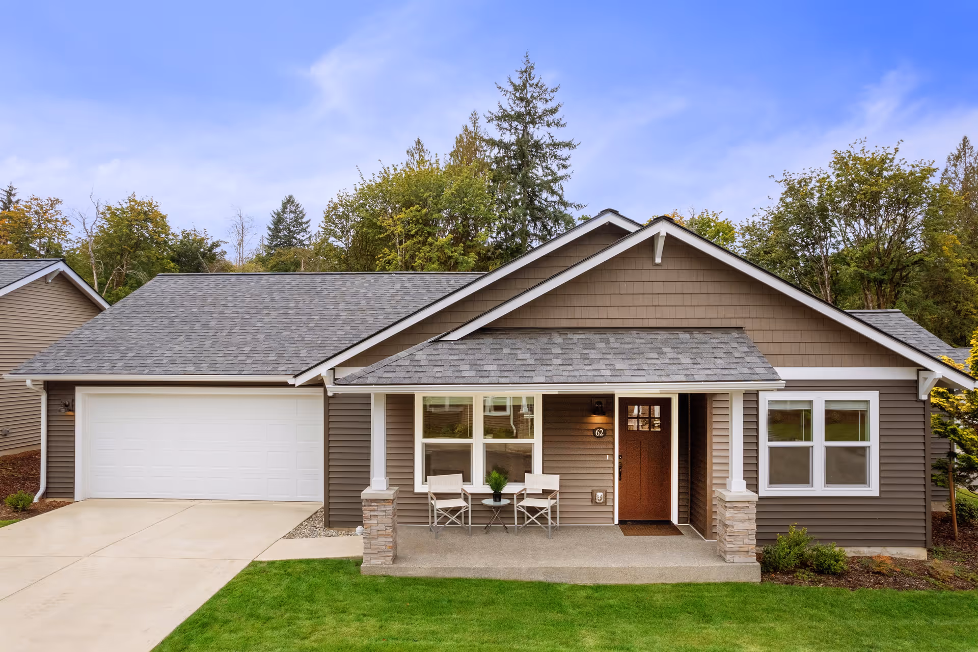 Front exterior view of a single-story house with a gray shingled roof, brown siding, white trim, a two-car garage, and a small covered porch with two chairs and a small table. The house is surrounded by green grass and trees in the background under a blue sky.